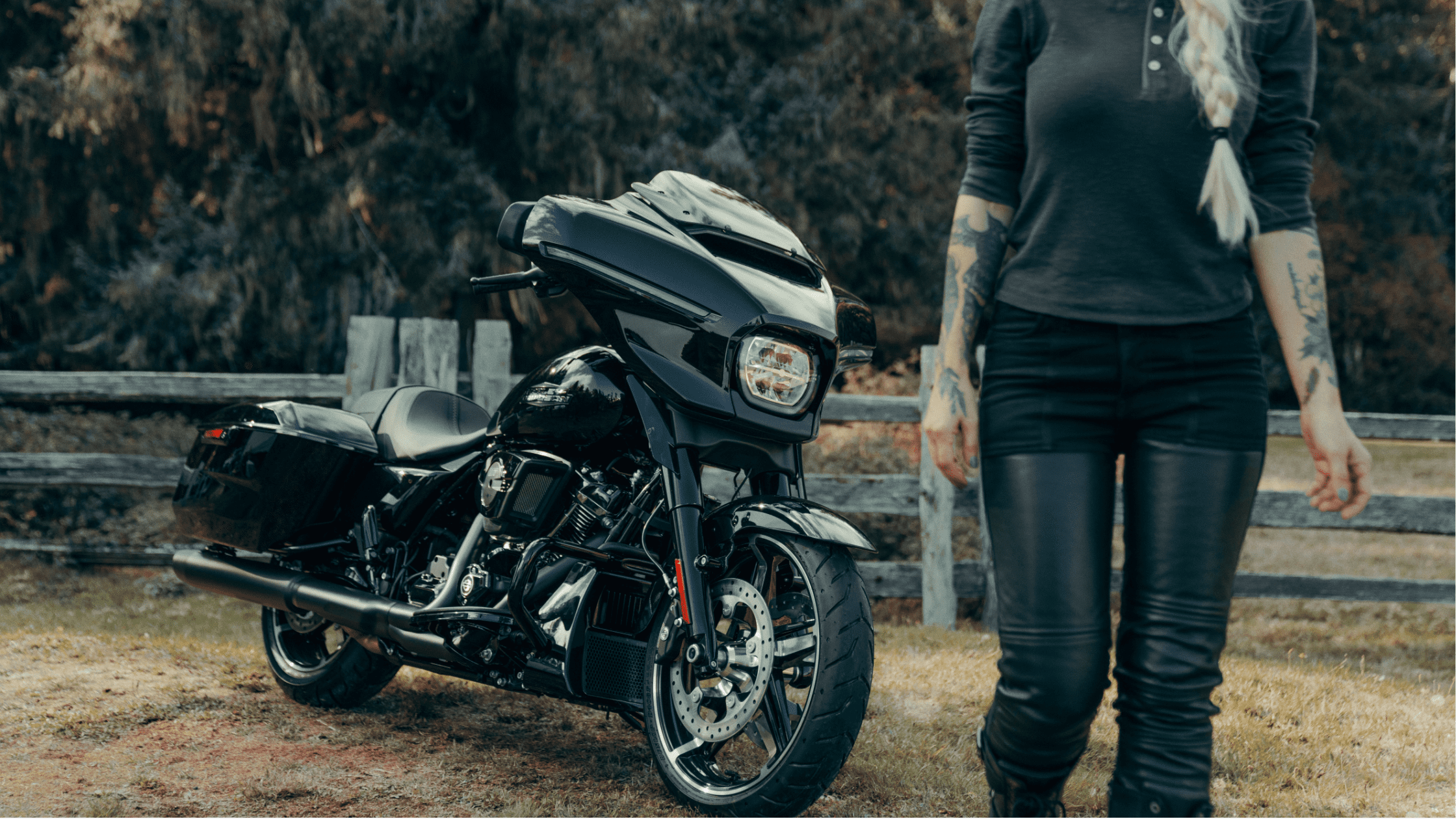 A sleek black 2024 Harley-Davidson® Street Glide® parked in a grassy area near a wooden fence, with…
