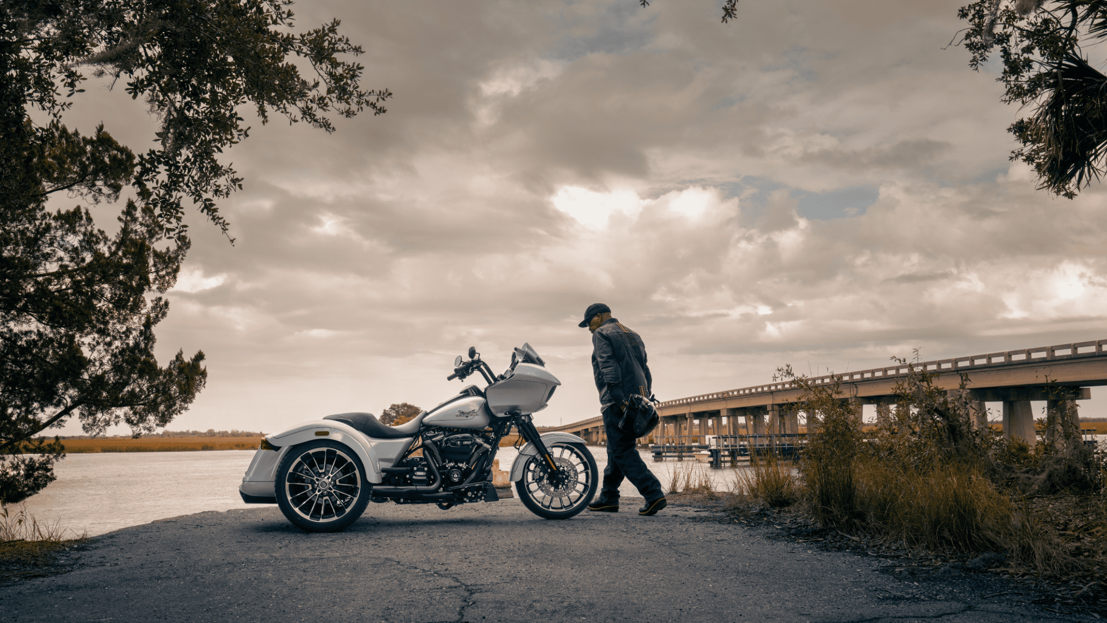 2025 Harley-Davidson® Road Glide® 3 trike parked near waterfront bridge under cloudy sky with rider…