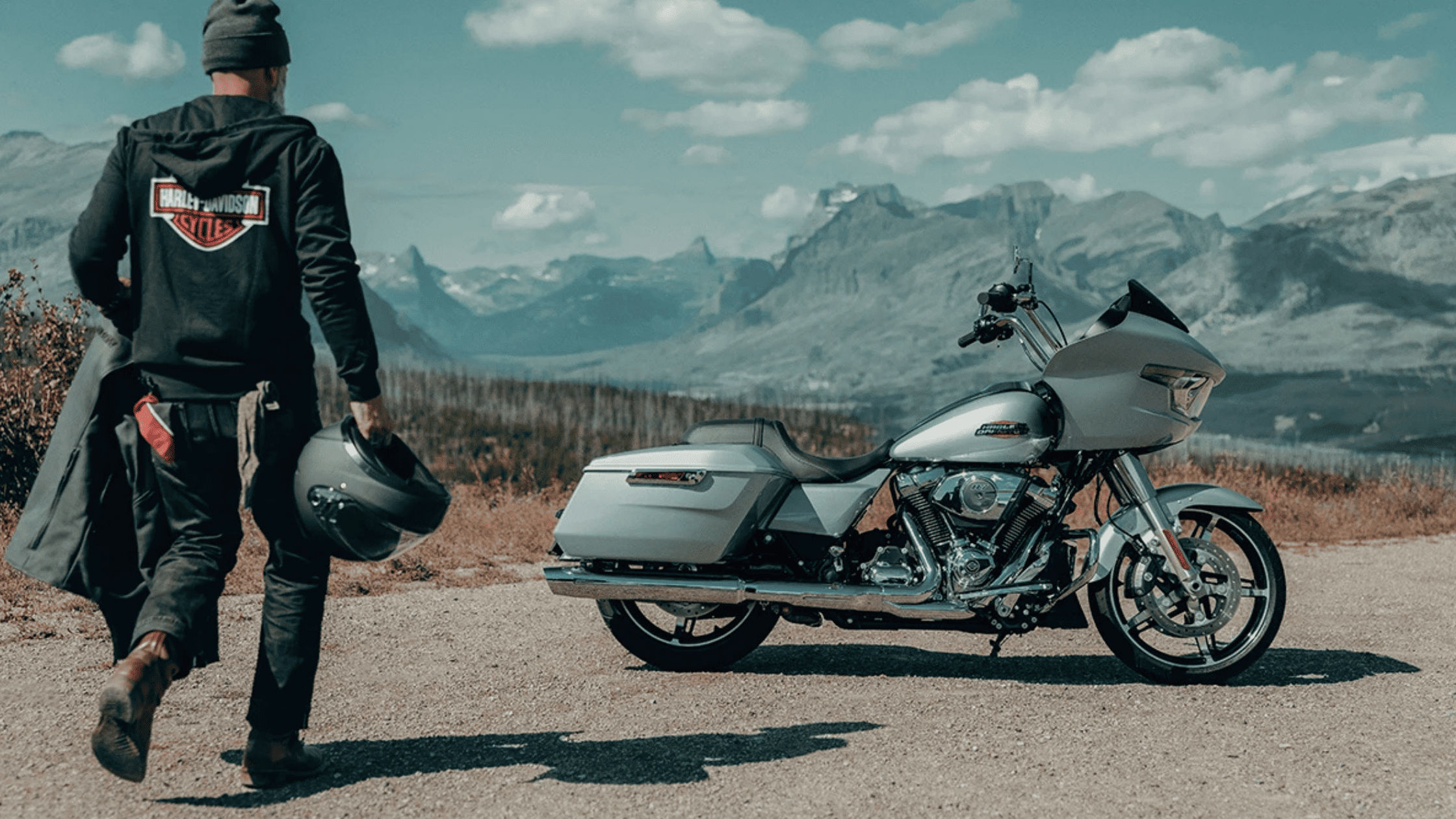 A rider approaching a silver 2025 Harley-Davidson® Road Glide® parked on a scenic mountain road